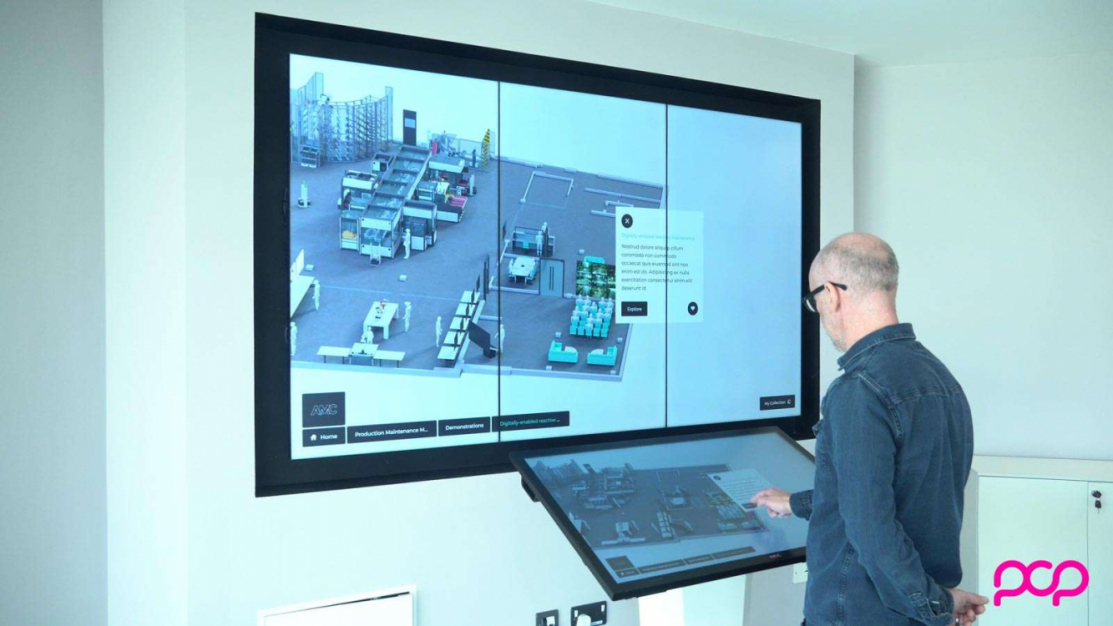 Interactive video wall displaying a 3d image of an advanced engineering factory which is controlled from a touchscreen with a man standing in front