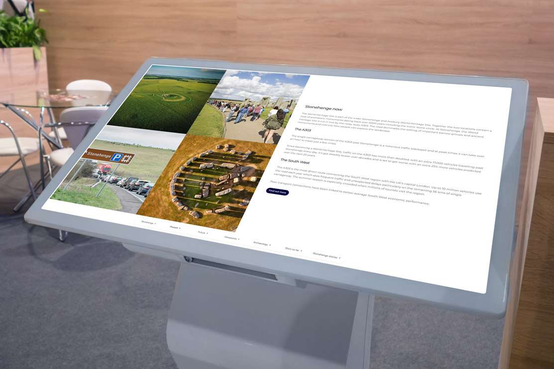 Large white interactive touchscreen in a meeting room with images of stonehenge, traffic and visitors, glass table in the background and chairs