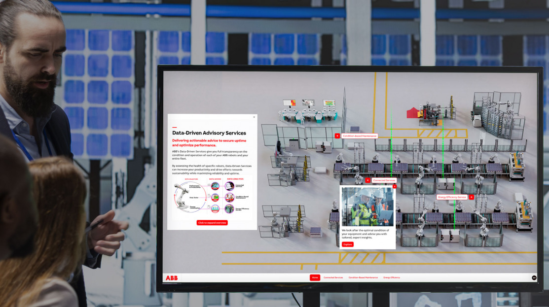 man standing by touchscreen talking to colleagues about a complex industrial factory floor with robots on the screen