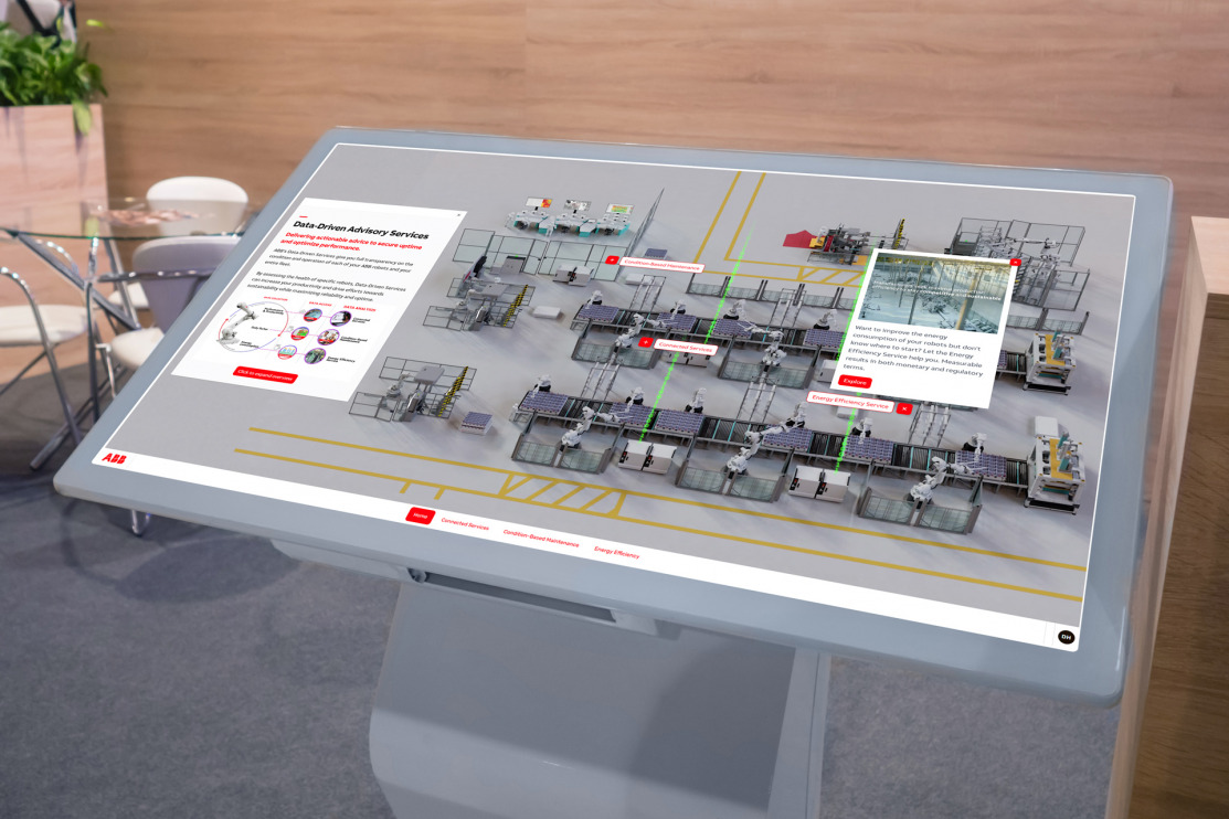grey touchscreen in foreground showing robots on a factory floor with text explanation on top, table and chairs in the background of booth for meetings