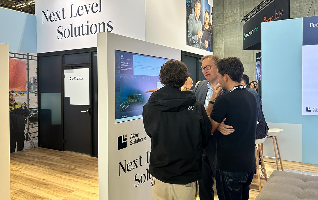 Three trade show visitors in conversation beside an Aker Solutions touchscreen kiosk, one gesturing towards a subsea equipment page on the screen.